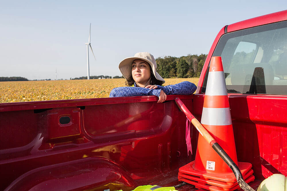 http://A%20person%20wearing%20a%20wide-brimmed%20hat%20leans%20on%20the%20side%20of%20a%20red%20pickup%20truck%20bed,%20looking%20into%20the%20distance.%20The%20truck%20bed%20contains%20traffic%20cones%20and%20a%20shovel.%20A%20wind%20turbine%20and%20a%20field%20are%20visible%20in%20the%20background%20under%20a%20clear%20sky.